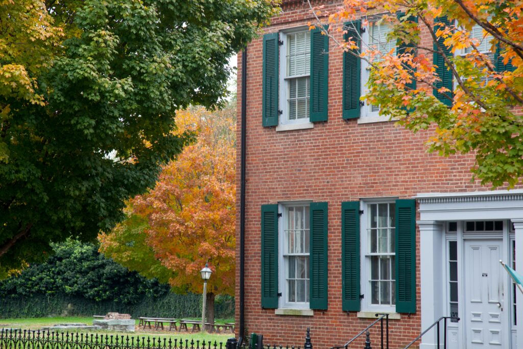 A brick building with green shutters is framed by colorful trees in a vibrant autumn scene.